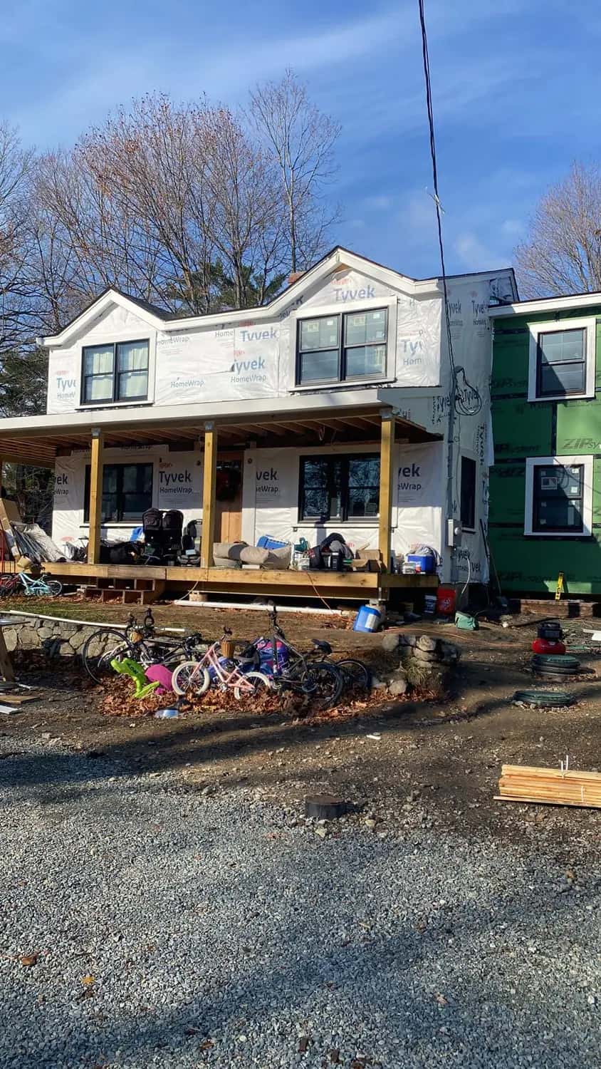 Original house in white house wrap with dormers and a new front porch, next to a two-story addition in green sheathing